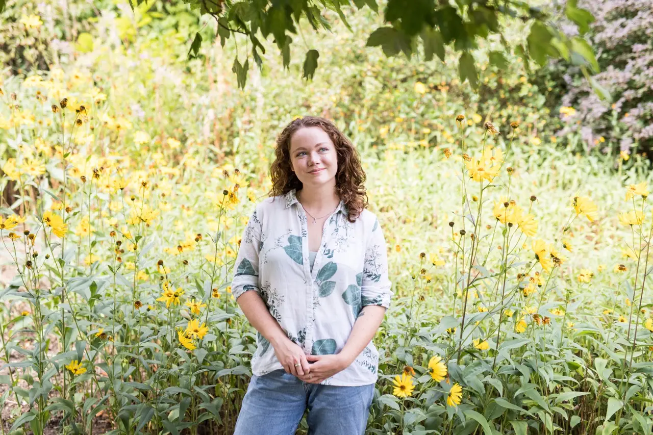Rebecca is wearing a white shirt and stop smiling in a field with yellow flowers and long grass behind her. 