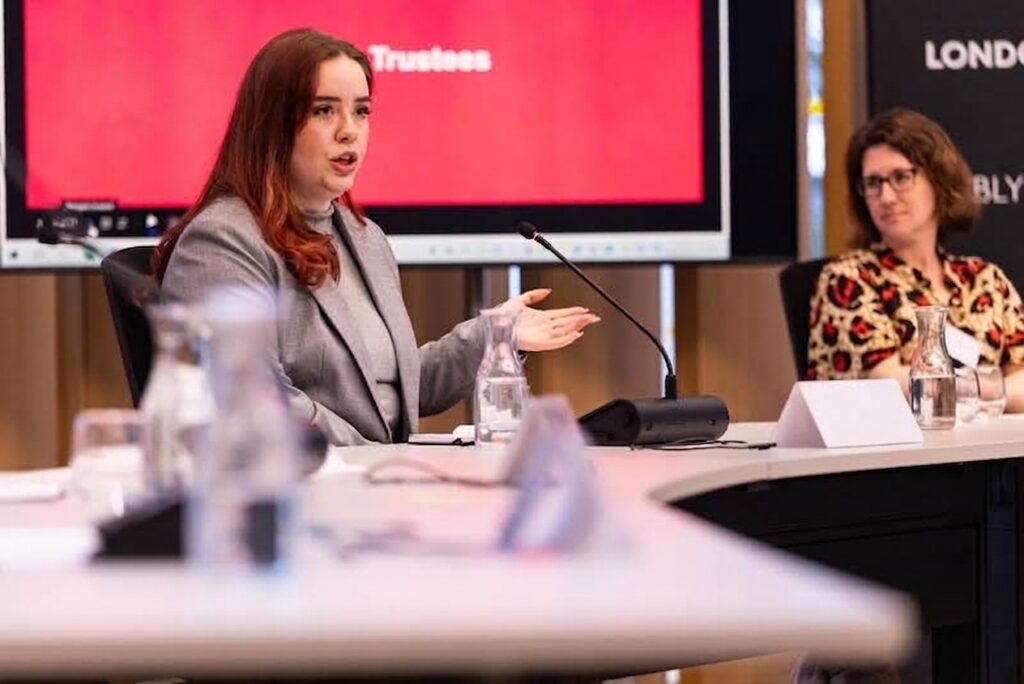 A young speaker presents at a panel discussion in front of a red screen, with another attendee listening nearby.